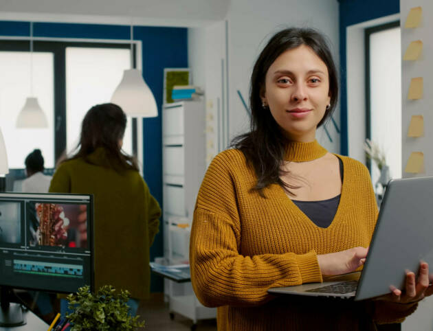 Woman retoucher looking at camera smiling working in creative media agency Woman retoucher looking at camera smiling working in creative media agency standing in front of webcam holding laptop. Photographer graphic editor working in start up office digital assets.